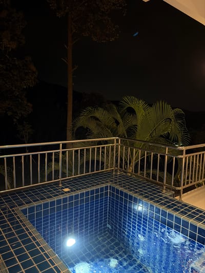 An outdoor swimming pool is illuminated by underwater lights at night. The pool is tiled with dark blue squares and is surrounded by a tiled deck. A stainless steel railing separates the deck from the surrounding area. Beyond the railing, palm trees and other foliage are visible against a dark night sky. In the far background, the silhouette of a hill can be seen. The location is specified as Ban Kao Dua, Thailand.