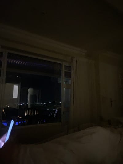 This image depicts a dimly lit interior room at night, located within the Ndlambe Local Municipality, South Africa. A large, white-framed window occupies the left side of the frame, providing a view of a dark cityscape illuminated by numerous scattered artificial lights in the distance. Immediately beyond the window, an outdoor area, possibly a balcony or patio, contains dark furniture, likely tables and chairs, with faint blue light discernible beneath them. Through the window, a tall, light-colored rectangular structure and another bright rectangular light source are visible. In the lower left foreground, a human hand holds a smartphone with an illuminated white screen. The room's interior is sparsely lit, primarily by the phone's screen and ambient light from outside. A rumpled bed, covered with white or light-colored sheets, occupies the lower right portion of the frame. The wall on the right side of the room is light-colored and features a dark door handle. Light-colored curtains are partially visible on the right side of the window frame, and crown molding runs along the top wall-ceiling junction.