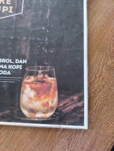 The image shows a promotional poster or advertisement placed on a brown woodgrain surface. The poster is printed with a background of dark grey to black, featuring a stylized depiction of a glass of iced coffee. The coffee beverage appears to be mixed with ice, milk, and coffee, creating a swirling effect of brown and white colors inside the glass. Small drops of a liquid are displayed above the glass, seemingly in the process of being added to the drink. The text on the poster includes partial words such as "RE UPI," "BROL, DAN MA KOPI," and "ODA," suggesting that the poster advertises a specific type of coffee or a coffee establishment. The image is likely related to the Indonesian city of Matang, and it is used to advertise a local coffee shop or beverage offering.