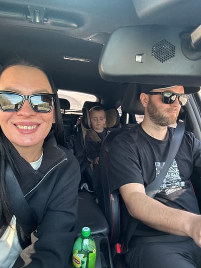The image displays the interior of a modern passenger vehicle, likely a car, occupied by three individuals during daylight. In the left foreground, an adult female with long dark hair wears a black zippered jacket over a white top and dark-framed sunglasses that reflect a road with guardrails. She is smiling and looking directly at the camera. In the right foreground, an adult male with a beard wears a dark baseball cap, dark-framed sunglasses, and a black t-shirt with a light-colored graphic print. He appears to be driving, looking forward. Both front occupants are secured by seatbelts. A young child, appearing to be a boy aged approximately 3-6, is seated in a forward-facing child safety seat in the rear passenger compartment. His eyes are closed, suggesting he is asleep or resting. A green plastic bottle of Lipton Iced Tea is visible in a cup holder on the center console between the front seats. The vehicle's interior features a dark headliner and dark seats with red accent stitching. A rearview mirror is positioned above the driver, displaying a hexagonal speaker grille and a small sensor. The exterior environment visible through the windows and in reflections indicates a road or highway with barriers under bright, clear conditions. The specific location within the city of Krivelevo, Russia, cannot be determined from the visual information present in the image.