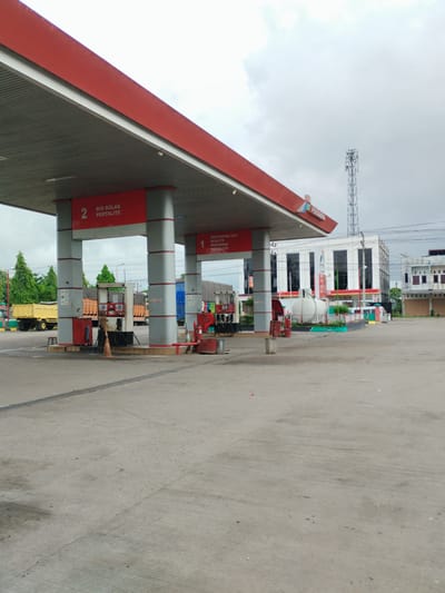 The image depicts a Pertamina gas station in the city of Payameuneng, Indonesia. The scene is dominated by the red and grey canopy of the gas station, with two prominent pillars labeled "2 BIO SOLAR PERTALITE" and "1 PERTAMINA DEX PERTAMAX PERTALITE". Several fuel pumps are visible under the canopy, with some trucks parked in the background. A white tanker truck is also visible, along with trees and buildings in the distance. The sky is overcast, suggesting a cloudy day. The ground is paved with concrete, showing some wear and tear. The overall impression is that of a functional, operational gas station.