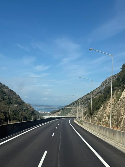 The image depicts a multi-lane asphalt highway, viewed from a forward perspective, likely from a moving vehicle, in the vicinity of Αγία Μαρίνα, Greece. The highway features two lanes in each direction, marked by solid white lines on the shoulders and dashed white lines separating the lanes. No other vehicles or individuals are present.
The road is flanked by steep hillsides. The left side is densely covered with dark green deciduous trees and vegetation. The right side features a rocky, sparsely vegetated incline with sections of retaining structures or netting. A series of uniform gray streetlights on tall poles extends along both sides of the highway, positioned behind concrete barriers that line the road.
In the mid-distance, the highway appears to traverse an elevated structure. Beyond this, a wide body of water, possibly a bay or gulf, is visible, bordered by landmasses and a cluster of light-colored buildings along the distant shore.
The upper portion of the scene is dominated by a clear, bright blue sky with faint, wispy white cirrus clouds scattered across it. The lighting suggests daytime conditions.