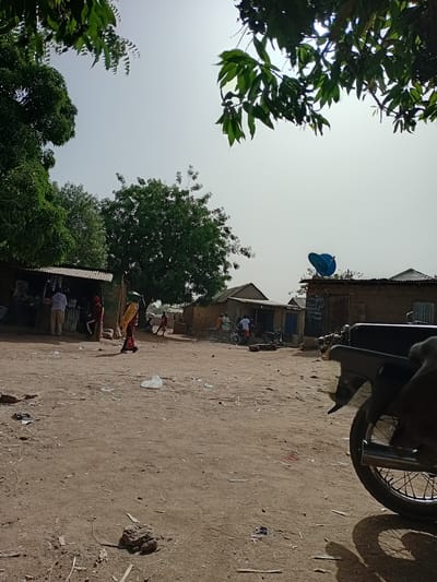 The image depicts a dirt road scene in Nata-Ala, Nigeria. The road is lined with buildings, some appearing to be shops or stalls. Several people are present, engaging in activities near the shops. A motorcycle is partially visible in the foreground. Trees and vegetation frame the scene, with a clear sky above. A blue satellite dish is mounted on one of the buildings.