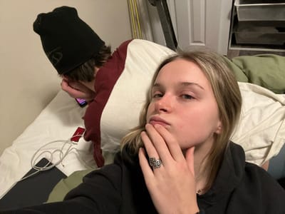 The image captures an indoor scene, likely a bedroom, featuring two individuals relaxing on what appears to be a bed. The perspective is from slightly above and angled, giving a candid feel. The overall lighting suggests an indoor setting, possibly daytime with curtains drawn or evening.

In the foreground, a person with light brown or blonde hair, styled with some darker undertones, is lying on their back or side on a white pillow. Their left hand is raised, partially covering the lower part of their face, including their mouth and chin. A distinctive, ornate silver-colored ring with a branch-like or abstract floral design is visible on their ring finger. Their visible eye appears open, looking towards the left side of the frame. The person is wearing a dark-colored, possibly black, long-sleeved top or hoodie. A green blanket or sheet is visible beneath them on the bed.

Behind and to the right of the first person, a second individual is partially visible, primarily their head and upper body. This person has short, dark brown hair and is wearing a black beanie with a subtle, lighter-colored graphic or logo on it. Their right hand is visible, holding a smartphone with its screen illuminated, suggesting they are actively using it. They appear to be lying on a maroon or deep red pillow.

The bed is covered with white and green bedding. On the white bedding, near the second person, there are white cables, possibly charging cables for electronic devices, along with a small, flat red object, which could be a power bank or a small case.

In the bottom left corner of the image, parts of household furniture are visible: a white door and a dark gray, multi-tiered storage unit or set of drawers. No discernible text is visible on any objects or surfaces in the image. The scene conveys a casual and relaxed atmosphere.