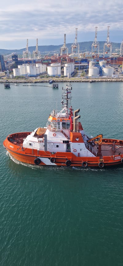An aerial view shows a bright orange and white tugboat named "ARES KOPER" moving through calm, green water in the port of Ankaran, Slovenia. The tugboat has a white superstructure with orange accents, a red crane, and several black fenders along its side. The tugboat is moving left to right across the frame. In the background, a large industrial port complex is visible, featuring numerous large white storage tanks, shipping containers stacked high, and several tall cranes. The sky above is partly cloudy.