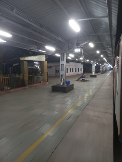 The image depicts an empty train platform in Igatpuri, India, at night. Overhead lights illuminate the platform, which is long and narrow. A yellow safety line runs along the edge of the platform. There are waiting areas with benches. The platform is under a metal roof. The perspective suggests the photo was taken from a train car.