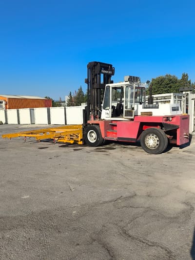 A forklift truck with a red and white cab and a yellow trailer attachment is parked on an asphalt surface. The forklift has "Kalmar LMV" and "ISOBOX" branding visible on its body. In the background, there are several modular buildings, some orange and some white, and trees under a clear blue sky. The scene is outdoors, likely in an industrial or storage area, possibly within the Municipal Unit of Inofyta, Greece, given the context of industrial facilities in the region. There are no people visible in the image.
