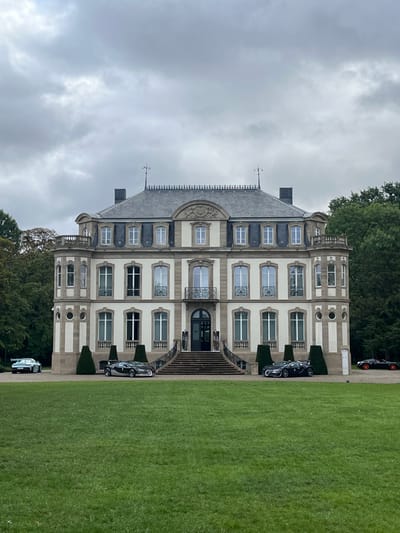 The image displays the Château Saint-Jean, located in Dorlisheim, France, under an overcast sky. The prominent three-story manor house exhibits a symmetrical facade of light-colored stone or render, accented with darker trim. A steep, dark grey mansard roof with numerous dormer windows and an ornate railing along its peak crowns the structure. A central grand entrance features a double staircase leading to a dark doorway, flanked by wrought iron railings, with a decorative balcony directly above. Conical topiary bushes are strategically placed along the paved perimeter of the building. An expansive, well-maintained green lawn occupies the foreground. Multiple luxury vehicles are parked on the paved area: a light blue Porsche is positioned on the far left, two dark grey Bugatti hypercars are situated closer to the building on either side of the main entrance, and a black Bugatti with orange accents is partially visible on the far right. Dense green foliage and trees border the background behind the château. No individuals are present within the scene.