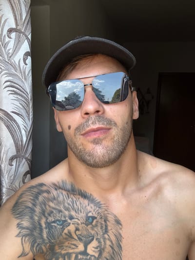 The image is a close-up, horizontally oriented, selfie-style photo of a person from the chest up. The person is wearing sunglasses and a dark hat. A large tattoo of a lion's face is visible on their chest and shoulder, featuring blue eyes. The background is dark, suggesting indoor lighting, possibly with some natural light coming from the side, reflected in the sunglasses, showing trees and sky. The person has stubble and a mole on their cheek. The lower part of the image shows a textured fabric, possibly a sofa or bedding, with a floral pattern in neutral tones. The overall mood is casual and posed. There is no visible text in the image. The context of Sesto Calende, Italy is provided, but there are no direct visual cues within the image to confirm this specific location.