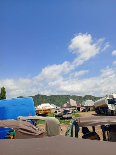 The image depicts an outdoor scene in Barakin Babele, Nigeria, under a clear blue sky dotted with fluffy white clouds. The time of day appears to be late morning or early afternoon, with bright sunlight casting shadows.
In the foreground, several auto-rickshaws, also known as "keke napep," are parked. Their brown fabric canopies are visible, with the interior of one showing a person wearing a striped shirt. Further back, a large blue vehicle, possibly a bus or truck, is parked on the left side of the frame.
The mid-ground reveals a collection of other vehicles, including a yellow truck, a white van, and a silver car. Beyond the vehicles, several buildings with distinct roofing styles are visible, suggesting a semi-urban or village setting. A large white tanker truck is parked on the right side of the image. In the background, rolling green hills rise, covered in vegetation, with some rocky outcrops.
The overall impression is one of a bustling but relaxed day. The weather is fair and sunny, conducive for outdoor activities and travel. The presence of various modes of transport and buildings indicates a populated area. There is no visible text in the image.