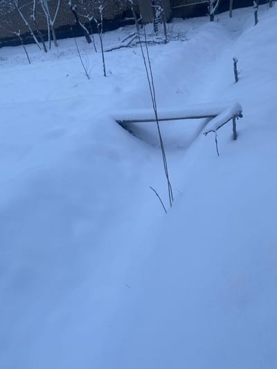 A winter scene shows a snow-covered landscape. Bare tree branches and thin wires emerge from the deep snow. A brick structure is visible on the left side, also coated in snow. The image is taken at an angle, giving a unique perspective of the snow's depth and the sparse objects sticking out from beneath it. The color palette consists mostly of white and blue, creating a cold, tranquil atmosphere.