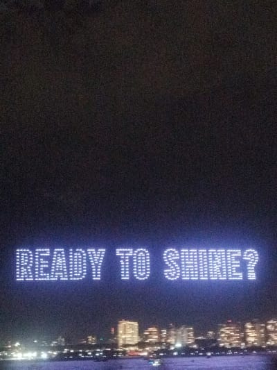 A nighttime sky displays the words "READY TO SHINE?" formed by numerous small, bright lights. Below the text, a blurred cityscape is visible, suggesting the message is displayed above a town or city. The sky is dark and unclear. The lights forming the words are a clear, bright white.