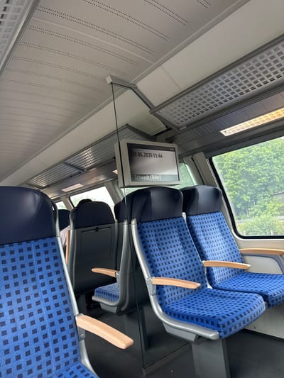 The image shows the interior of a train carriage. In the foreground, there are two seats upholstered in blue fabric with a black checkered pattern. The seats have grey metal frames and light-colored wooden armrests. To the left, another identical seat is partially visible. Further back in the carriage, more seats can be seen, as well as a person seated facing away from the camera. A screen is mounted on the ceiling, displaying the date "29.06.2026 11:44" and the text "Schwedt (Oder)". The ceiling is white with a perforated pattern, and a fluorescent light fixture is visible. Through the window on the right, lush green trees and foliage are visible, indicating an outdoor environment. The overall scene depicts a typical train interior, likely in transit. The location in Trebbin, Germany, is not directly evident from the visual information presented in the image, but the text on the screen suggests a journey to or from Schwedt (Oder).