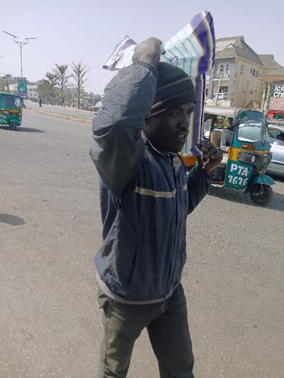 The image depicts a male individual walking on a paved street in Mumbwa, Zambia, under clear daylight conditions. The subject, of dark complexion, occupies the foreground, wearing a dark knit cap, a dark blue zip-up jacket with two light horizontal stripes across the chest, and dark trousers. He carries a blue, white, and brown patterned textile bundle balanced on his head, secured by his raised hands. His mouth is slightly open.
In the midground, to the left, a green auto-rickshaw is partially visible, oriented away from the camera. To the right, another green and yellow auto-rickshaw, bearing the alphanumeric "PTA 7676", is present, with a silver vehicle partially visible behind it. The background features a multi-story building with a multi-gabled roof and visible scaffolding, indicating ongoing construction or renovation. Solar-powered streetlights and palm trees line the street. A billboard is partially visible on the left side of the road. The paved surface appears to be asphalt or concrete.
