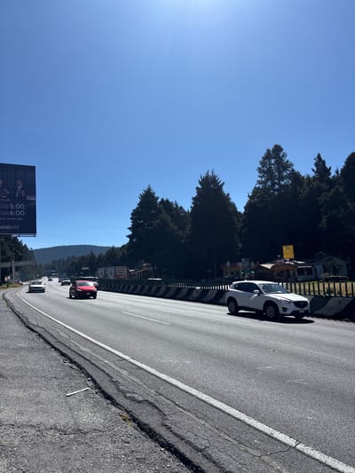The image, which appears to be rotated 90 degrees clockwise from its natural orientation, depicts a scenic roadside view on a bright, sunny day. A wide asphalt road, featuring a clear white dividing line and a slightly cracked shoulder, occupies the lower right portion of the frame. A black and white striped barrier or guardrail runs along the side of the road, separating it from the adjacent terrain.

To the upper right of the road, a dense forest of dark green coniferous trees lines the landscape, extending upwards towards the top of the frame. Beyond this immediate treeline, in the mid-distance on the upper left, more forested hills or mountains are visible under a clear, azure sky. The sky itself dominates the upper left and central parts of the image, showing a brilliant blue with a subtle sun flare or light ray visible in the top left corner, suggesting strong daylight conditions.

Several vehicles are present on and near the road. A white car, possibly a compact SUV or sedan, is parked on the side of the road closest to the camera, facing away. Further down the road, a red car is visible, followed by several other vehicles receding into the distance, indicating traffic flow or parked cars.

Behind the white parked car and partially obscured by the trees, some small buildings or stalls with various commercial signs are discernable, suggesting roadside establishments.

In the lower left corner of the image, a large billboard stands prominently. Visible text on the billboard includes "las 5:00.", "las 6:00.", and "EN UN SOLO LUGAR". A partial brand name, "AND MX", can also be seen. The billboard features images of people, though specific details are unclear.

The overall impression is one of a clear, warm day, likely in a mountainous or hilly region, with a road winding through a lush forest.