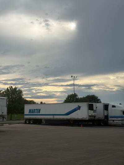 This is a vertical, eye-level, medium shot of a cloudy sky over a parking lot in Murfreesboro, United States of America. The sky is filled with a mix of dark gray and light gray clouds. A patch of bright white light shines through the clouds in the upper right quadrant of the image. In the middle ground, a white semi-trailer truck with a blue stripe and the word "MARTEN" in blue lettering is parked. The truck has two axles and a tandem axle. The cab of the truck is white with a blue stripe. The truck is parked on asphalt. In the background, there are green trees and a lamppost with three lights. A small orange traffic cone is on the ground in front of the trailer.