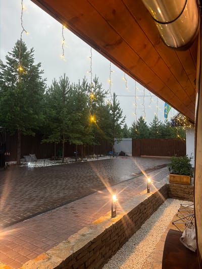 The image depicts an outdoor paved area, likely a courtyard, with a wooden overhang extending into the frame from the upper right. The ground is paved with reddish-brown interlocking bricks and appears wet, suggesting recent rain. A metal drain grate runs across the paving. Along the edge of the paved area, a low wall constructed of stone and brick is visible. This wall is illuminated by two modern, rectangular, solar-powered pathway lights, each emitting a warm yellow glow. Beyond the wall, a narrow strip of white gravel is present.
A line of trees, predominantly pine trees, with dense green foliage, forms a backdrop to the scene, extending across the width of the image. Behind the trees, a dark wooden fence encloses the property. In the distance, a lighter colored metal fence is visible. The sky is overcast and a pale grey, indicating a cloudy day or dusk.
Decorative strings of small, warm white fairy lights are draped from the wooden overhang, with some strands extending towards the trees. A flagpole with what appears to be the Ukrainian flag is visible on the right side of the image, attached to a building structure. A planter box containing green foliage sits on top of the stone and brick wall on the right. In the foreground, to the far right, a dark wooden surface, possibly a table or bench, is partially visible, with a folded grey blanket resting on it.
There are no people present in the image. The overall impression is one of a well-maintained outdoor space, possibly a residential property in a rural or suburban area, with decorative lighting suggesting an evening or early morning setting. The location is identified as Kolyutkino, Russia.