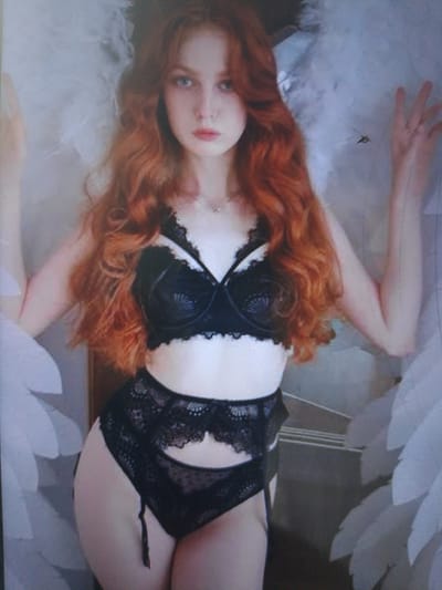 A young woman with long, wavy red hair and blue eyes is posing in lingerie. She is wearing a black lace bra and matching bottoms with suspenders. She also has large white feathered wings attached to her back. The background is softly lit and out of focus, with what appears to be a wall or large piece of furniture behind her. The image has a dreamy, ethereal quality. The woman's expression is calm and direct. There is no visible text in the image. The location context is Dobra Krynytsia, Ukraine, but there are no specific visual cues in the image to confirm this. The time of day and weather are not discernible.