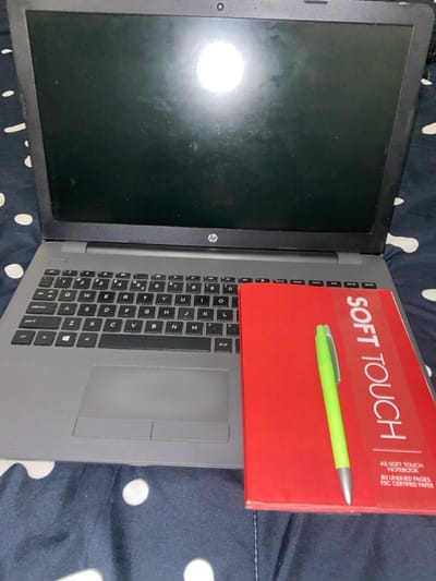 An HP laptop with a closed screen is partially covered by a red notebook labeled "SOFT TOUCH". A lime green pen rests atop the notebook. The laptop's keyboard and touchpad are visible. The background is a fabric with a dark blue and white polka dot pattern. The notebook is an A5 Soft Touch notebook with 80 unlined pages of FSC certified paper. The keys Q, W, E, R, T, Y, U, D, H, K, C, V, N, and M are visible on the keyboard.