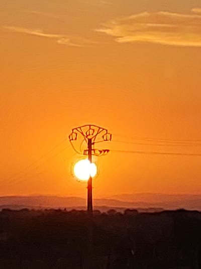 The image depicts a sunset scene with an electric pylon silhouetted against a bright orange sky. The pylon is centrally located and features a distinctive, ornate top structure. A bright white orb, likely the sun, is positioned behind the pylon, partially obscured by its structure and wires. In the foreground, a dark, silhouetted landscape with rolling hills is visible, hinting at the natural environment surrounding Sobradillo, Spain. The sky above the horizon is a gradient of orange and yellow, with faint wispy clouds. The overall impression is one of a serene and dramatic sunset in a rural or semi-rural setting.