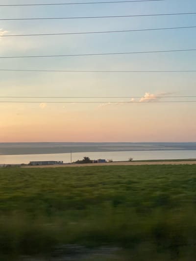 The image depicts a rural landscape at sunset, featuring a vast body of water and cultivated fields. The sky transitions from a pale blue at the top to hues of orange and pink near the horizon, with a few scattered clouds. Several parallel power lines stretch across the upper portion of the frame. In the middle ground, a body of water, likely a river or lake, separates the foreground from a distant shore. Along the edge of the water, several structures are visible, including what appear to be modular buildings or shipping containers, along with some vegetation and a solitary pole. The foreground is dominated by a large, grassy field, appearing slightly blurred due to motion, suggesting the photo was taken from a moving vehicle. The scene is devoid of human presence. The overall impression is one of tranquility and natural beauty at the end of the day.
