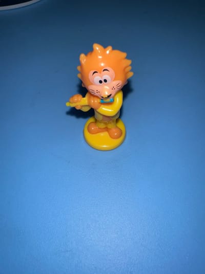 The image displays a small, colorful plastic figurine positioned horizontally on a plain blue surface. The figurine depicts a cheerful, cartoon-style character, likely an animal, with a predominantly orange head and a yellow body and base. Its head features spiky, fur-like orange details and a friendly facial expression with large white eyes, black pupils, and a small, smiling mouth. Tiny black lines on its cheek suggest whiskers. A light blue element, possibly a bow tie or collar, is visible around its neck. The figurine's body is yellow, and it stands securely on a round, flat yellow base.
The background is a solid, royal blue surface with a subtle texture, consistent with an indoor setting like a table or floor. The lighting appears to be artificial, casting soft shadows beneath the figurine, particularly around its yellow base. No specific activity is taking place; the figurine is stationary. There is no visible text in the image. The orientation of the image shows the figurine lying on its side, relative to its upright position.