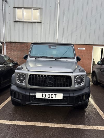 A silver-grey Mercedes-Benz G-Class SUV, displaying UK registration "13 OCT" with "CAR COMPONENTS" text below it, is parked head-on in a commercial or industrial area within Nibley, United Kingdom. The vehicle, appearing damp from moisture, is situated on a wet asphalt surface within a marked parking bay. Its front features a black grille with a black Mercedes-Benz emblem and circular LED headlights. To the left, a dark-colored sedan is partially visible. To the right, a light-grey taxi with "EURO TAXI" lettering is partially visible. The background consists of a building with a lower section of reddish-brown brickwork and an upper section of light-grey corrugated metal siding. A white-framed window is present in the upper section, and a doorway marked with a white "DIRECTOR" sign is visible within the brick section on the right. Overcast lighting conditions are apparent.