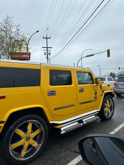 The image features a striking, extended yellow Hummer H2 SUT (Sport Utility Truck), likely a limousine, stopped on a wet road in Pitt Meadows, Canada. The vehicle is highly customized with large, aftermarket wheels featuring yellow spokes and chrome rims, as well as chrome running boards and door handles. Water droplets cover its surface, indicating recent rain.

The perspective is from inside another vehicle, with its dark side mirror partially visible in the foreground on the right, reflecting parts of the road and the yellow Hummer.

The setting appears to be a commercial area or intersection. In the background, other vehicles are visible, including a white SUV directly behind the Hummer. Overhead, numerous power lines stretch across the frame, supported by utility poles. A traffic light and a street light are also present. To the left, a large, prominent red sign with the golden arches and "McDonald's" text is visible, partially obscured by a tree with budding or bare branches. The sky is overcast, contributing to the muted lighting. Visible text includes "H2" and "SUT" badging on the Hummer's side and "McDonald's" on the restaurant sign.