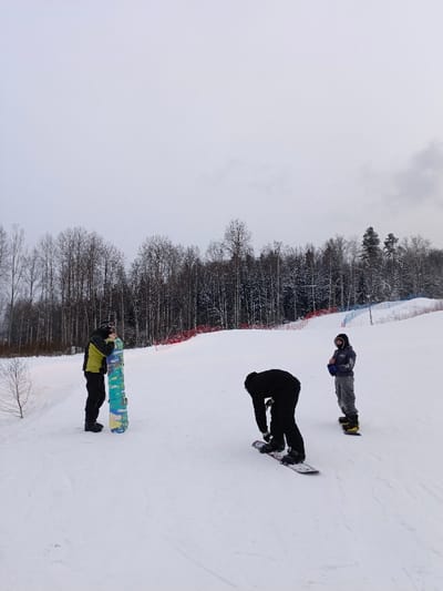 The image captures a winter scene on a snow-covered slope, likely a ski resort or snowboarding park, under an overcast sky. Three individuals are present on the white expanse, engaged with their snowboards.
In the foreground to the left, a person is standing upright, holding a snowboard vertically in front of them. This individual is dressed in a vibrant lime green and black jacket, black pants, and appears to be wearing a dark helmet or balaclava. The snowboard itself features a distinctive camouflage-like pattern in shades of light blue, yellow, and green.
Slightly to the right and in the mid-ground, another person is bent over, facing away from the viewer. This individual is entirely clad in black attire, including a jacket and pants, and is focused on adjusting the bindings of their snowboard, which is lying flat on the snow beneath them.
Further to the right and slightly behind the person in black, a third individual stands upright on their snowboard. This person is looking towards the left side of the frame and is wearing a grey hooded jacket, grey pants, and a dark helmet or goggles. Their snowboard is primarily yellow with black accents.
The background features a dense forest of trees, many of which are deciduous and bare, though some evergreen trees are also visible, all lightly dusted with snow. Running along the edge of the slope, behind the people, are safety nets or fencing, distinguished by sections of red and blue, indicating boundaries or hazard areas. The sky is a uniform, pale grey, suggesting a cloudy winter day without direct sunlight. There is no visible text in the image. The overall impression is one of a recreational winter outing, with the individuals preparing for or taking a break from snowboarding.