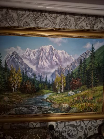 This is a painting of a mountainous landscape. The scene features snow-capped mountains in the background, with a lush green forest and a winding river in the foreground. There are several trees with yellow and brown foliage, suggesting it might be autumn. The river flows over rocks, and the surrounding landscape is covered in grass and small pink flowers. The sky is a clear blue with a few wispy clouds. The painting is framed in an ornate gold frame. There are no people visible in the painting. The overall mood is serene and natural. The image is a vertical shot, and the painting is hanging on a wall with patterned wallpaper.