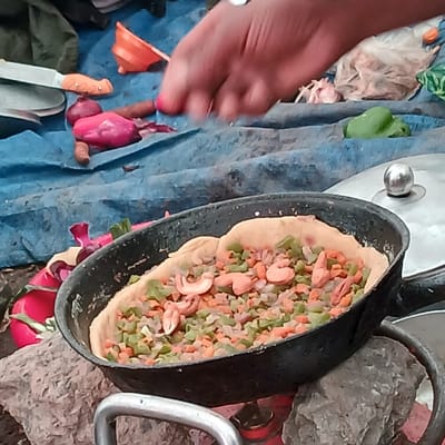 In Gilman's Point, Tanzania, a close-up, elevated view captures a person preparing food outdoors. The central subject is a black, oval-shaped pan containing a light-colored dough base topped with a mixture of finely diced green vegetables (likely bell pepper or zucchini), orange vegetables (likely carrots), and reddish-purple pieces (likely red onion). Several whole or half cashew nuts are interspersed throughout the topping. The pan rests on a makeshift cooking setup, supported by grey rocks or bricks, with a red gas cylinder partially visible beneath.
In the upper portion of the frame, a light-skinned hand with short, painted fingernails (red/pink polish) is positioned over the cooking area, seemingly in the process of adding ingredients or manipulating the food. The immediate surface surrounding the pan is covered by a wrinkled blue tarp or fabric. Scattered on this blue surface are various raw ingredients: two whole red onions, a small pile of peeled carrots, a green bell pepper, and several cloves of garlic partially contained within a light-colored mesh bag. Other objects visible include a small orange funnel, a metal knife with a light handle, and the edge of a silver pot lid with a spherical knob. The overall scene suggests food preparation in a rustic or informal setting.