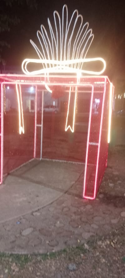 A large, illuminated gift box decoration stands on a paved surface. The box is outlined with red lights, with white light ribbons and bow on top. The gift's interior is a shaded space. A street with a white car is visible through the box. The paved ground features irregular stone patterns, and grassy areas with fallen leaves surround the decoration. The overall ambiance is nighttime.