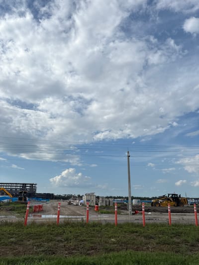 The image depicts a construction site in Palmetto, Florida, United States, under a partly cloudy sky. In the foreground, there is a grassy area with dry soil patches. A row of orange and white traffic cones marks a boundary. Behind the cones, a dirt path leads into the construction site, with several puddles of water visible.
Further into the site, construction materials are scattered. To the left, a large, multi-story building structure is under construction. Various construction equipment, including a yellow excavator, is present. There are stacks of construction materials, possibly concrete pipes and other building components. A white portable restroom or storage unit is also visible.
In the mid-ground, a tall concrete utility pole stands in the center, with multiple power lines running horizontally across the frame. To the right of the pole, a yellow Caterpillar motor grader is actively working on the uneven terrain. More construction materials and partially built structures are visible in the background, along with trees and houses in the distance. The overall scene suggests an active construction project in progress.