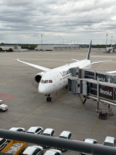 The image shows a Lufthansa Boeing 787-9 Dreamliner parked at Munich Airport (Flughafen München, MUC) in Freising, Germany. The aircraft is connected to two jet bridges advertising "Revolut". A row of white cars are parked adjacent to the aircraft. The sky is cloudy. The overall scene appears to be taken from an elevated position within the airport terminal.