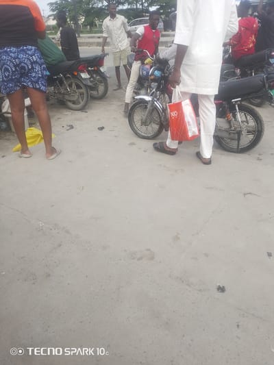 In Eputu Town, Nigeria, a group of people are gathered around motorcycles. A woman in the foreground wears a blue and white patterned skirt and a black and orange top. To her left, a man in a black shirt stands near a green tarpaulin covering part of a motorcycle. Further back, a man in a patterned white shirt and light shorts stands next to a motorcycle. To the right, another man in a red vest and white pants is near a motorcycle, and a person in a white outfit is holding a red and white shopping bag. Several motorcycles are visible, with one in the foreground having a license plate that is partially obscured. The ground is paved with concrete, and trees and buildings are visible in the background. The overall impression is a busy street scene with people preparing to travel or disembarking from motorcycles.