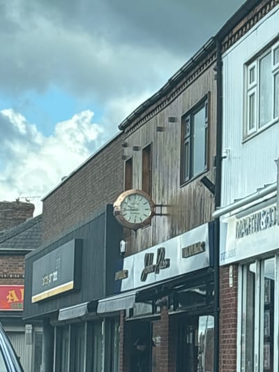 This image shows the exterior of the bar/restaurant "Tom's Tap" located in Crewe, United Kingdom. An outdoor clock hangs on the building's side, next to the sign. Above, a mix of brick and wooden paneling is visible. The sky is overcast with patchy clouds.