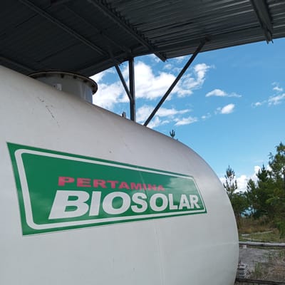 The image shows a close-up of a large, white cylindrical tank with a green and white logo that reads "PERTAMINA BIOSOLAR". The tank appears to be outdoors, under a corrugated metal roof supported by metal beams. The background consists of a bright blue sky with scattered white clouds and a line of trees, some of which appear to be pine trees. The scene suggests an industrial or storage facility. The specific location in Gegarang, Indonesia, is not directly visible in the image but is provided as context.