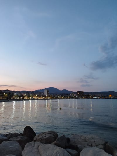 The image depicts a twilight view of the coastline in El Campello, Spain, looking towards the city. The sky transitions from a pale blue at the top to shades of pink and dusky blue near the horizon, with a few wispy clouds. The city lights are prominently reflected in the calm sea, creating a shimmering effect. Buildings, including a tall, modern tower and other structures, are visible along the shoreline, interspersed with palm trees. In the foreground, large, grey rocks are scattered along the edge of the water. A yellow buoy is visible in the sea. The overall scene is tranquil, capturing the early evening atmosphere of a coastal town.