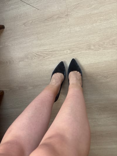 The image shows a top-down view of a person's legs and feet, wearing pointed-toe black shoes, on a light-colored wooden floor. The legs are bare and tanned. The scene is indoors, likely a residential setting, with no indication of the city of Prodanovtsi, Bulgaria. The wooden floor has visible grain and some minor scuffs or scratches.