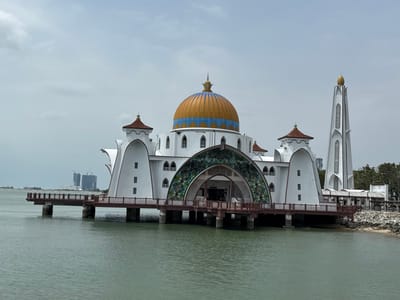 The Malacca Straits Mosque sits on stilts above the water. Its central dome is gold with blue accents, flanked by white arches and a matching minaret. The mosque's facade features an arched entrance adorned with intricate green and brown patterns. A walkway extends from the mosque towards the open water on one side, while the mosque connects to a sandy shoreline on the other. Distant buildings are visible on the horizon. The overall scene is bright under a cloudy sky.