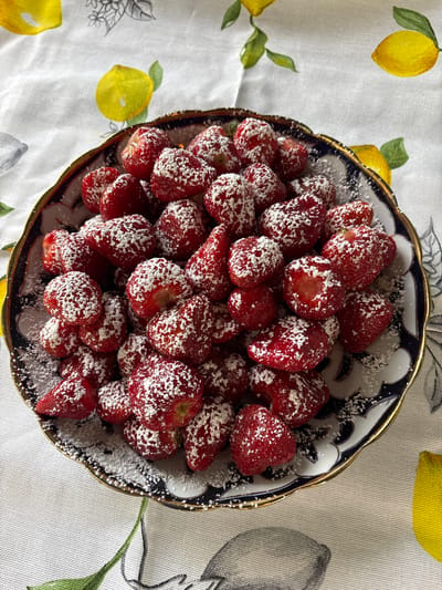 A close-up, overhead shot showcases a dark, ornate bowl filled with ripe strawberries dusted with powdered sugar. The strawberries are a vibrant red, with some appearing slightly translucent, revealing their juicy interior. The powdered sugar is unevenly distributed, creating a frosty, appealing texture. The bowl itself has a decorative pattern of dark blue and white, bordered by a gold rim. It rests on a light-colored tablecloth adorned with a pattern of yellow lemons and green leaves. The overall scene is bright and cheerful, suggesting a delicious treat prepared for enjoyment. There are no people visible, and no text is present in the image. The context of "Пермяки, Russia" suggests this might be a home setting or a local establishment in that region, likely during daytime given the lighting.