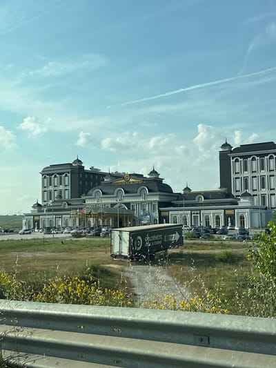The image captures the Goldeneye Hotel & Casino in Kapitan Andreevo, Bulgaria, from a roadside perspective on a clear day with scattered clouds. In the foreground, a grey metal guardrail runs horizontally across the bottom, behind which is an unkempt grassy field with prominent yellow flowering plants and tall dry grasses. A dirt path cuts through this vegetation, leading towards the casino complex. A dark-colored semi-trailer truck, bearing the "GOLDENEYE HOTEL & CASINO" logo on its side, is positioned on this path in the mid-ground.
The casino complex is a large, multi-story structure exhibiting ornate, classical architectural styling. It features dark grey domed roofs with gold accents and light-colored facades. The main central section of the building prominently displays "GOLDENEYE CASINO" signage near its roofline. To the left and right, taller, rectangular wings with multiple windows and decorative elements flank the central structure. A grand covered entrance with columns is visible in the central section, with what appear to be flags flying nearby. A large paved parking lot in front of the casino is occupied by numerous parked cars. In the far left background, beyond the casino, power lines and pylons are faintly visible against the horizon. The sky is bright blue with scattered white cumulus clouds and several faint white contrails from aircraft. No individuals are clearly discernible in the image.