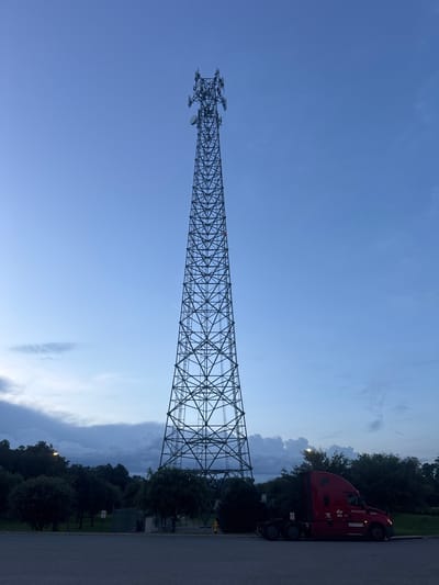 A tall, lattice cellphone tower stands prominently in the center of the image, dominating the skyline under a twilight sky. The tower is constructed of dark metal beams forming a complex geometric pattern. Attached to the upper sections of the tower are various antennas and equipment. To the right of the tower, a red semi-truck cab is parked. The truck features a sleek design and displays the company name "CR England" and the number "240445" on its side. The truck is parked on an asphalt surface, which appears to be a parking lot or roadside. In the background, a line of trees with dark green foliage creates a natural border. The sky transitions from a deep blue at the top to lighter shades near the horizon, with scattered clouds adding texture to the scene. The overall impression is one of an industrial and transportation hub against a natural backdrop, likely in the outskirts of a city.