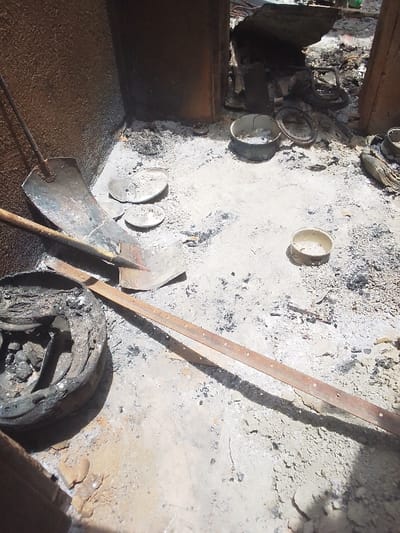 The image shows the aftermath of a fire in a building located at latitude 9.804, longitude 8.646 in the city of Kwal, Nigeria. The scene is a single room, presumably a kitchen or storage space. The floor is covered in ash and debris, indicating a recent fire. Several metal objects are scattered around the room including two metal shovels, two small round metal containers, a large blackened object in the foreground which may be a tire or a metal drum, and a long rusty metal bar. The walls of the room are made of brown brick and concrete and appear to have been scorched by fire. There is no indication of people being present in the room. The room is poorly lit but appears to be in the daytime, based on the shadows of the debris.