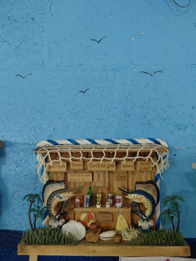 The image depicts a miniature, three-dimensional diorama of a coastal bar setting. Key features include wooden signage indicating offerings such as "Beer," "Wine," "Sailfish Bar," "Bud Light," and "Bacardi." The bar is adorned with miniature bottles and cans, along with nautical decorative elements such as marlin fish figurines, seashells, and artificial palm trees and foliage. The structure of the bar appears to be made of wood, wrapped with nautical rope and netting. The entire scene is placed against a textured, light blue surface, possibly representing the ocean or sky.
The location of Cutler Bay, United States of America is provided solely as context for the image and is not visually discernible within the image itself.