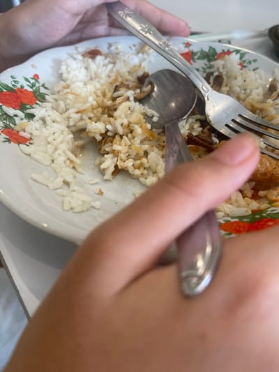 In Medang, Indonesia, a close-up shot captures a partially eaten plate of white rice with visible pieces of meat and fried onions. A fork and spoon rest on the plate, partially obscured by hands, suggesting a meal in progress. The plate itself has a floral pattern with red and green accents. The lighting is bright, indicating an indoor setting with natural light.