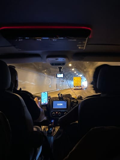 The image is taken from inside a vehicle traveling through a tunnel, possibly at night. The tunnel walls are illuminated with lights. In the foreground, we see the heads and shoulders of at least two individuals. One individual is driving, with their hands on the steering wheel and a smartphone mounted on the dashboard showing a navigation app. Another individual is seated in the passenger seat, their silhouette visible. A dashcam is mounted on the windshield. In the distance, a yellow truck is visible, along with other vehicles and tunnel lighting. The vehicle has interior lighting, including a red LED strip along the roof. The overall impression is that of a journey through an urban tunnel. The city of Gomilsko, Slovenia is mentioned in the prompt but cannot be confirmed from the visual information alone.