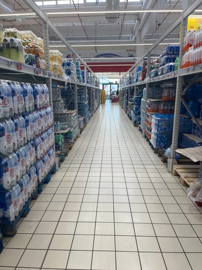 The image shows the interior of a supermarket aisle in Castiglione delle Stiviere, Italy, with shelves stocked with bottled water and other beverages. The flooring is tiled in a light color with dark grout lines. The aisle is wide and appears well-lit, with overhead fluorescent lighting. On the left side of the aisle, tall shelves are filled with cases of bottled water, many of which are shrink-wrapped. Some of the bottles have labels visible, with prominent brands and colors. On the right side, similar shelving units are also stocked with cases of bottled water, as well as some other types of drinks in bottles and multipacks. Pallets are visible at the base of some shelves, holding additional stock. In the distance, at the end of the aisle, two individuals are partially visible near a shopping cart, one of whom appears to be a shopper and the other a supermarket employee. The environment is clean and organized, characteristic of a retail grocery store.