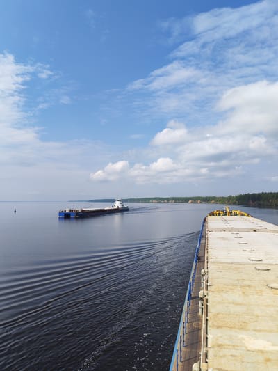 The image depicts a waterway near the village of Глебово, Russia. The view is from the deck of a barge, showing a section of the vessel's deck on the right side. A tugboat towing a barge is moving away in the distance on the left side of the frame. The water surface is dark, showing ripples and waves created by the movement of the barges. The sky is blue with scattered white clouds. A shoreline is visible in the background.
