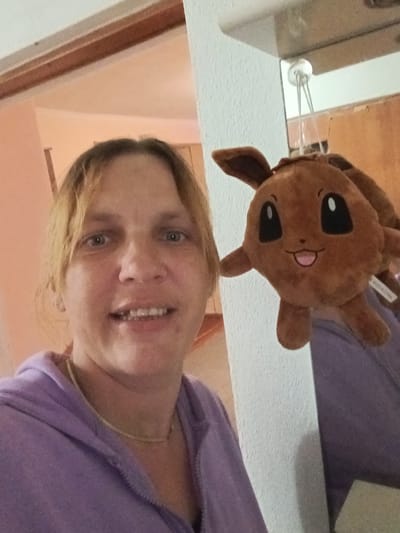 A middle-aged woman with blonde hair and blue eyes smiles at the camera while holding a brown stuffed Eevee toy. She is wearing a purple hooded jacket and a gold necklace. The woman is standing in what appears to be a hallway with a doorway to her left and a white textured wall to her right. The lighting is warm, suggesting an indoor setting. There is a reflection of the ceiling light fixture in a mirror or reflective surface on the wall. The background is blurry, but the general impression is of a domestic environment. There is no information in the image that specifies the location as Litynia, Ukraine.